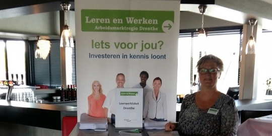 Adviseur  van het Leerwerkloket Drenthe poseert met de banner van het Leerwerkloket. Op de banner staat: Leren en Werken Arbeidsmarktregio Drenthe. Iets voor jou? Investeren in kennis loont.