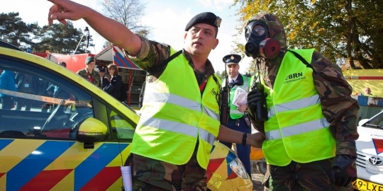 Twee medewerkers van defensie. Een heeft een gasmasker op en luistert naar de instructie.